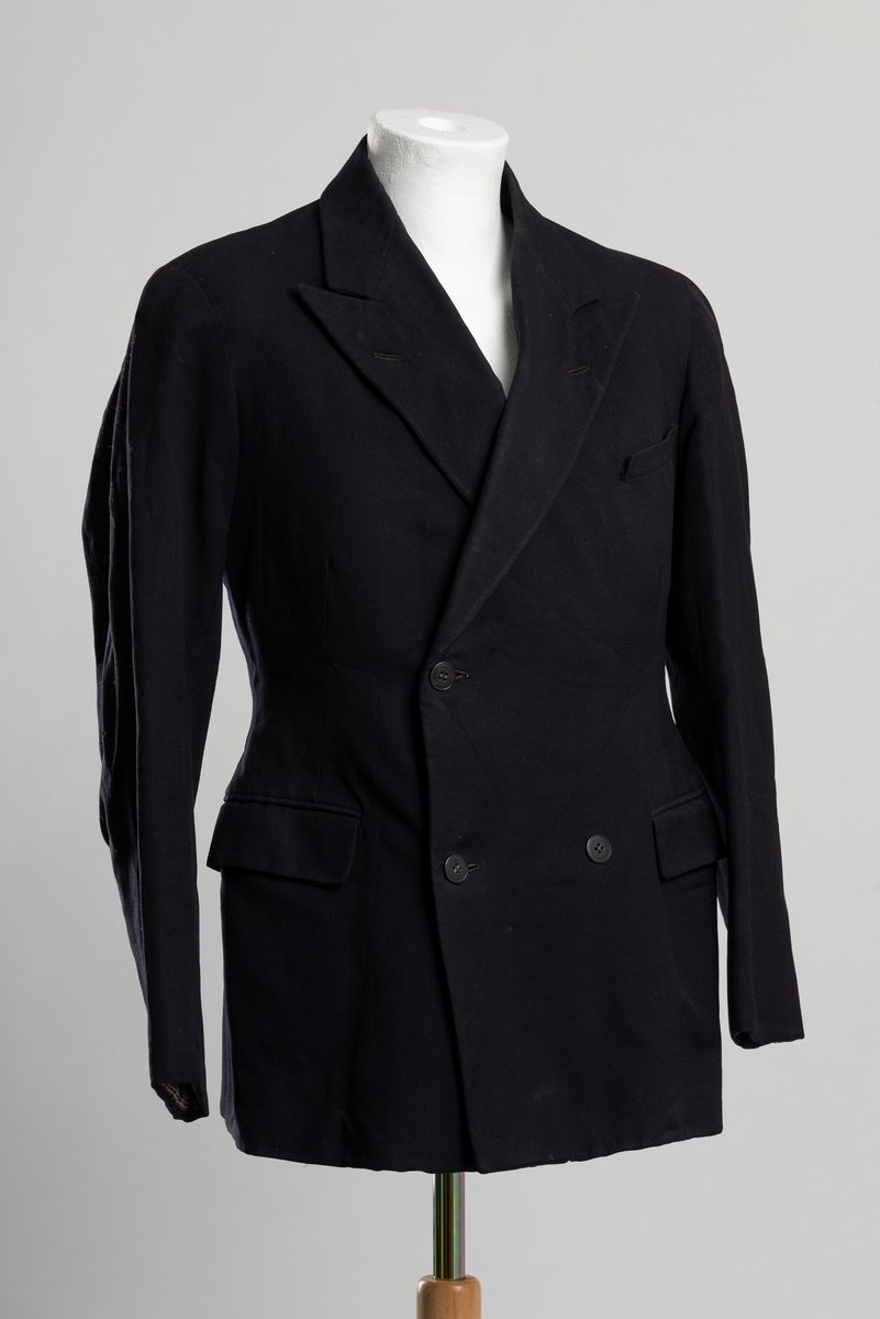 Svart dobbeltspent dressjakke. To lommer i front med klaffelukning og brystlomme på venstre side.  Innerlommer på begge sider. Knappelukning i front, fire knapper hvorav en mangler. Reparert i foret på høyre erme.