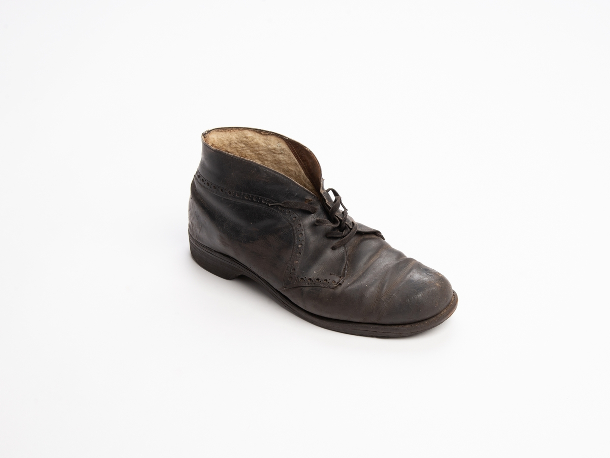 Ankelstøvel av svart lær med saueskinnsfor, høyrefot. Hullmønster langs sømmene på over siden. Snøring i front. Reparert på innersiden ved tåkappen. Løs innersåle. Satt inn ny innvendig hælkappe av saueskinn.