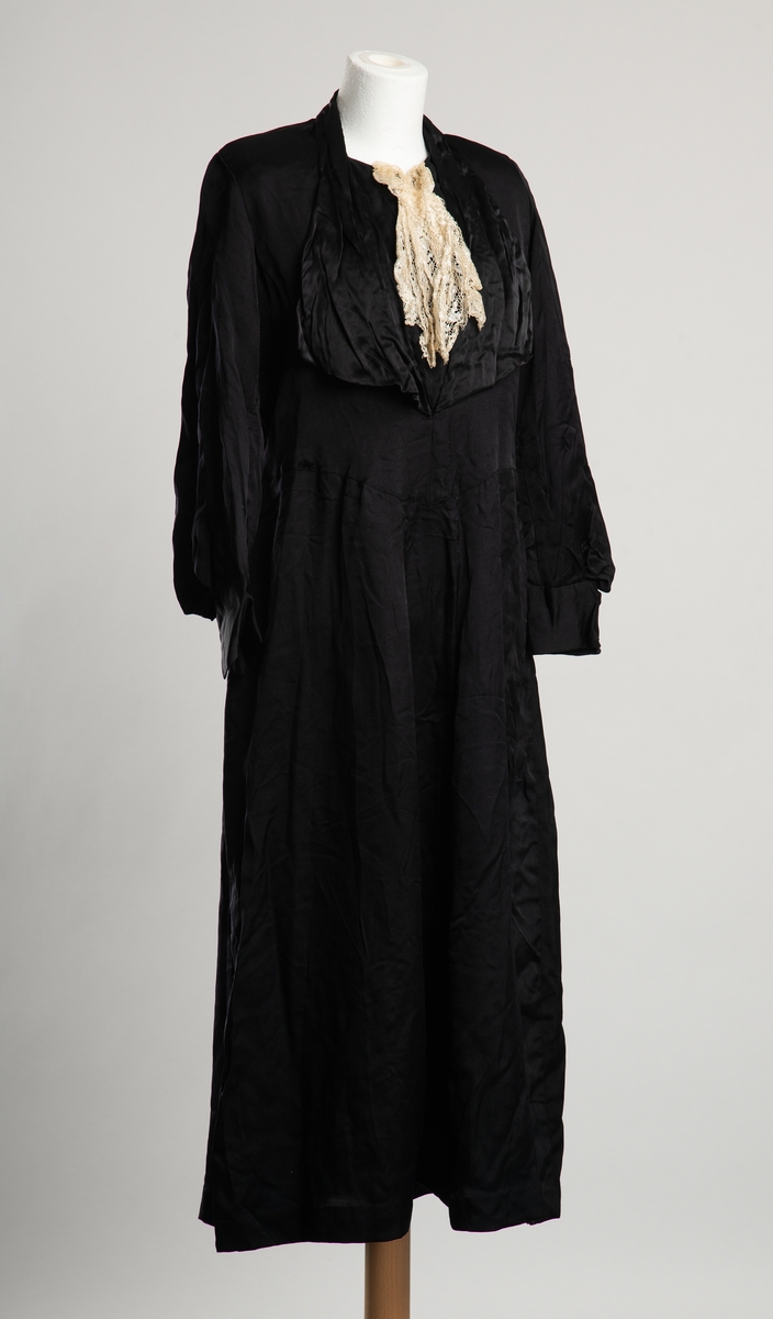 Fotsid kjole av svart sateng. Krage med store buede slag på brystet. Belegg i halsåpningen,  festes med trykknapp, dekortert med hvit blonde. Livet går ned i en spiss foran. Belte i ryggen med trykknapplukning på venstre side. Skulderputer, ballongermer med bred manskjett.