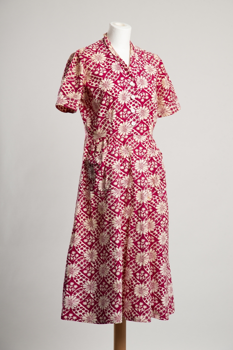 Knelang kortermet kjole med liten krage. Bomullskanvas med trykket blomstermønster. Åpning i overdel i front med knappelukkning, i tillegg glidelåsåpning i venstre side. To lommer på skjørt foran.