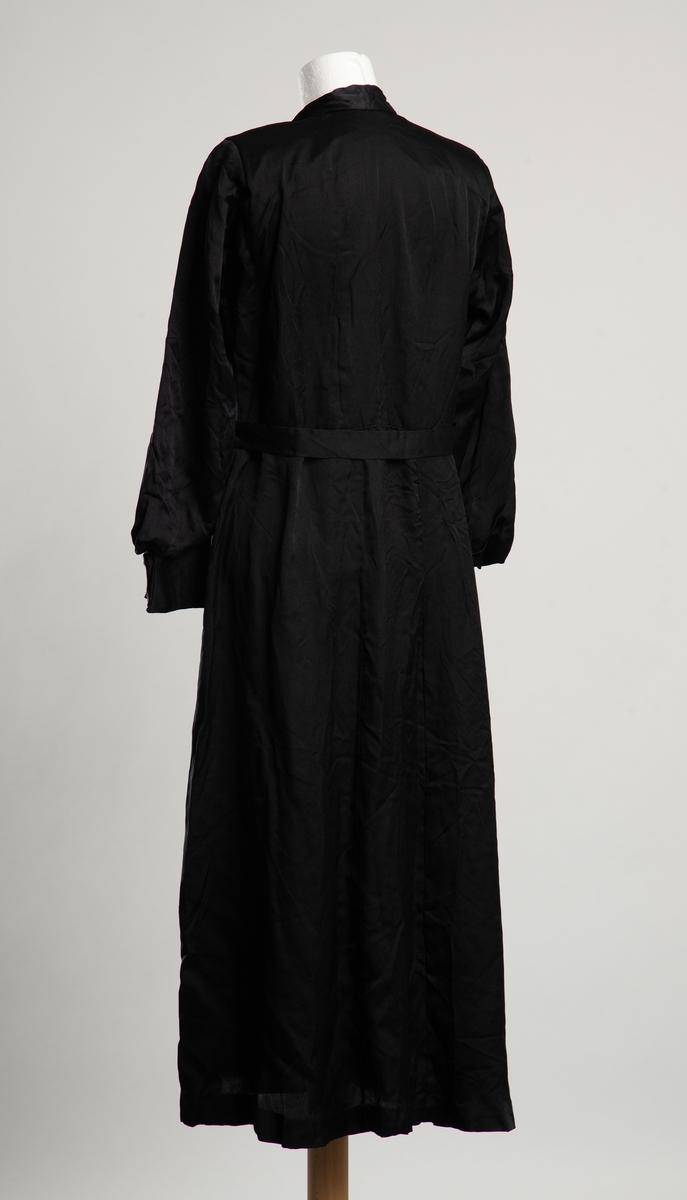 Fotsid kjole av svart sateng. Krage med store buede slag på brystet. Belegg i halsåpningen,  festes med trykknapp, dekortert med hvit blonde. Livet går ned i en spiss foran. Belte i ryggen med trykknapplukning på venstre side. Skulderputer, ballongermer med bred manskjett.