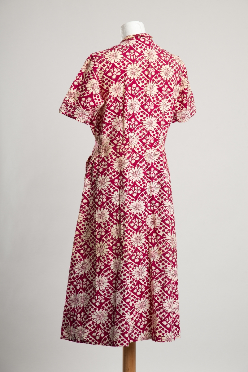 Knelang kortermet kjole med liten krage. Bomullskanvas med trykket blomstermønster. Åpning i overdel i front med knappelukkning, i tillegg glidelåsåpning i venstre side. To lommer på skjørt foran.