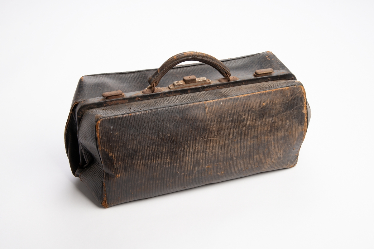 Rektangulær koffert av presset papp trukket med svart preget skinn. Åpning på langs med låsbar metallbøyle, nøkkel henger i snor. Hank av lær. Innvendig trukket med ubleket bomull. Sekundært innvendig trekk som er festet med sikkerhetsnåler. Inni kofferten ligger en pipette med emballasje, konvolutt med navn og en plastmappe.