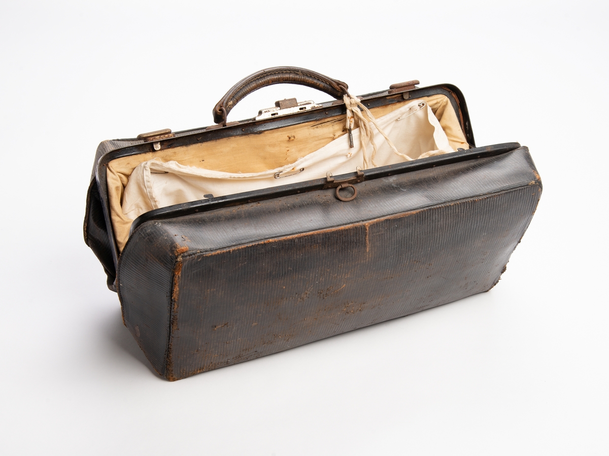 Rektangulær koffert av presset papp trukket med svart preget skinn. Åpning på langs med låsbar metallbøyle, nøkkel henger i snor. Hank av lær. Innvendig trukket med ubleket bomull. Sekundært innvendig trekk som er festet med sikkerhetsnåler. Inni kofferten ligger en pipette med emballasje, konvolutt med navn og en plastmappe.