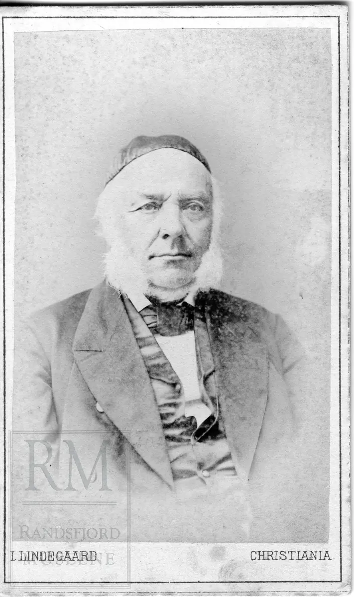 Brystbilde av mann med hvitt kinnskjegg. Han har en "pavelue" på hodet.