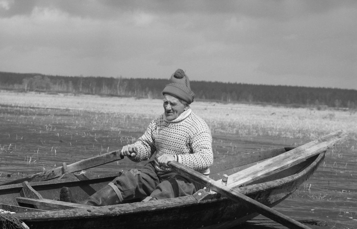 Joseph Bakken (1898-1989), fotografert i den klinkbygde robåten han brukte under utsetting av gjeddegarn i Tjønnan, et cirka 3 kvadratkilometer stort deltaområdet ved elva Tufsingas utløp i innsjøen Femund, i Tufsingdalen i Os kommune i Nord-Østerdalen. Joseph Bakken var kledd i vadmelsbukser og islender (strikkegenser med spettemøsnster). Han hadde ei strikket duskleue - topplue - på hodet og langskaftete gummistøvler på beina. Her skulle det settes garn for å fange gjedde, som i dette området, bortiimot 700 meter over havet, gyter like etter at isen har gått, ei ukes tid i mai måned. Det  er nettopp i denne perioden at det er enklest å fange såkalt «isgjedde», når den søker inn mot de grunne sivbevokste strendene for å gyte, Det var et slikt gyteområde Joseph Bakken rodde i da dette fotografiet ble tatt.