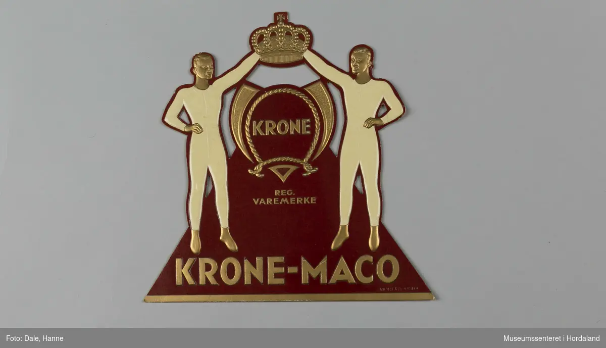 Reklame for Krone-Maco og Krone-merket frå Salhus Tricotagefabrik ved Bergen. Den er laga i påtrykt papp med "fot" på baksida slik at den kan stå på ein disk eller i ei hylle.