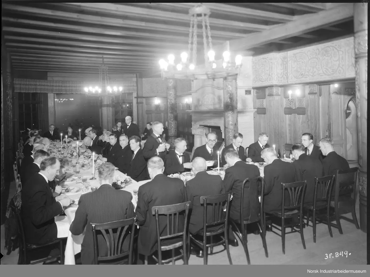 40 og 25 års jubilanter 1945. Jubilantene spiser middag i stuen på Admini. Mannlig servitør serverer drikke ved bordene.