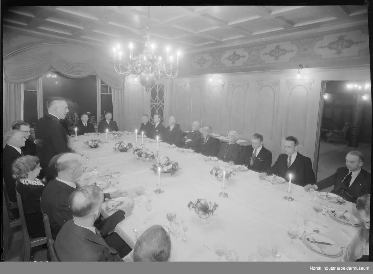 40 og 25 års. jubilanter 1945. Jubilantene sittende rundt dekket bord. Mann stående, holder muligens tale.