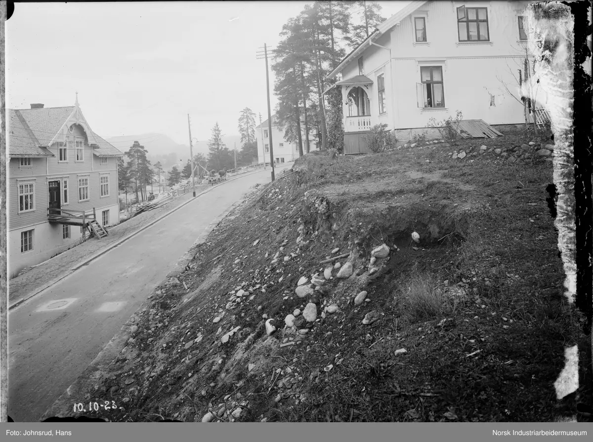Skråning ved vei under hus. Flere bygninger langs veien.