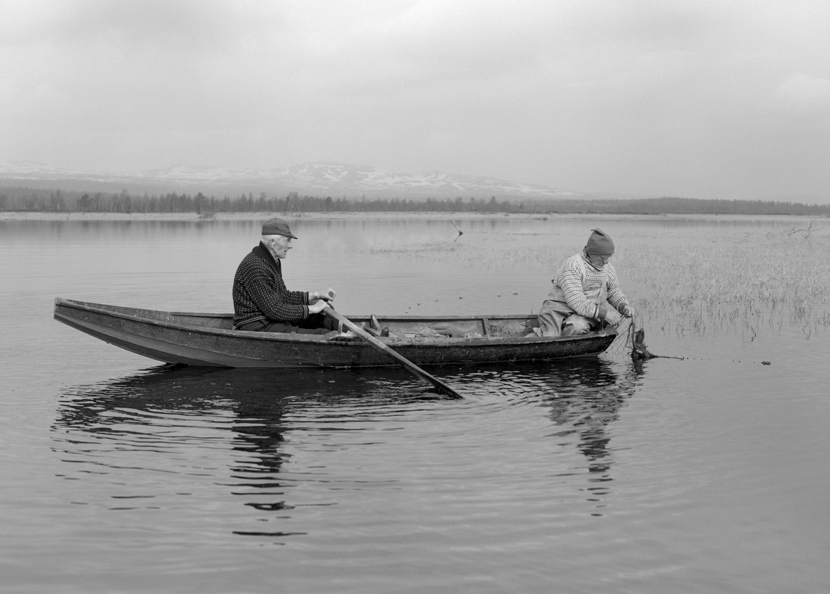 Jo Bakken (til venstre) og Jo Bakken fra Tufsingdalen i Os kommune i Nord-Østerdalen, fotografert under opptak av gjeddegarn fra en robåt i Tjønnan, det cirka 3 kvadratkilometer store deltaområdet der elva Tufsinga renner inn i innsjøen Femund. Gjeddefisket her starter like etter at isen har gått om våren. Da trekker gjedda inn mot de grunne, sivbevokste strandområdene for å gyte. Der fanges de med garn som settes skrått inn mot strandlinjene. Hvert garn er 35 meter langt, men stikker bare cirka 1 meter ned i vannet. Mer er ikke nødvendig, for garna skal stå på svært grunt vann. Dette garnfisket brukte å pågå cirka ei ukes tid, og i løpet av en slik periode fortalte Joseph Bakken at han kunne fange fra 100 til 300 kilo gjedde. De fleste av disse fiskene veide mellom 2 og 5 kilo. Joseph Bakken var kledd i islender og en overall av mer vannavstøtende stoff. Det sistnevnte plagget skulle antakelig forebygge at de klærne fiskeren hadde under ble søkkvåte av drypp fra det nyopptrukne garnet. Da dette fotografiet ble tatt var Joseph i ferd med å slå ihjel ei gjedde som satt i garnet ved hjelp av en krokete kjepp. Naboen Jo Bakken satt ved årene. Han hadde vadmelsbukser på beina, islender på overkroppen og hatt på hodet. De varme klærne var nødvendige, for det var ennå tidlig på våren og kaldt i lufta i Tufsingdalen.