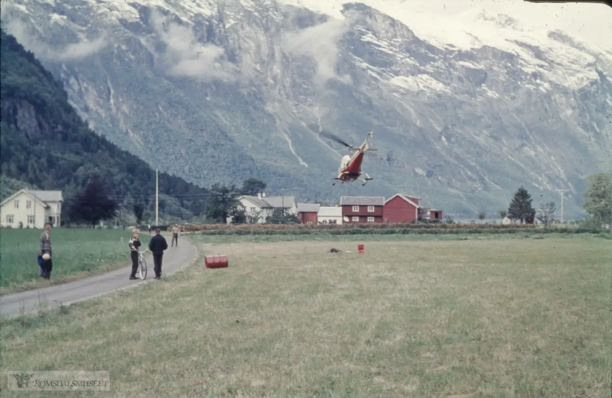 Reitan, Eikesdalen, gnr. 79/1. Helikopteret har truleg eit oppdrag for NVE knytt til Mardøla-utbygginga..LN-ORI er et Bell 47J Ranger helikopter tilhørende Helikopter Service..Husa er frå venstre Stuevoll, bnr. 36, bygd som kårhus på garden, fråskild nr. 1 i 1915. Våningshuset (kvitt) i to høgder. Framom dette står eit kastanjetre, no borte (2021). Våningshuset på bnr. 2 (raudt). Landhandelen eller «butikken» (i bruk frå 1905-1948) og eldhuset (kvitt i ei høgd), stabburet (bygd 1937-38) og t.h. for dette, fjøsen.