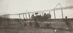 Flygplan SW 10 Farman med motor igång står på flygfältet på Malmen, cirka 1914-1915. Flygförare i flygplanet och personal ur Flygkompaniet i arbete bredvid. Vy bakifrån flygplanet.