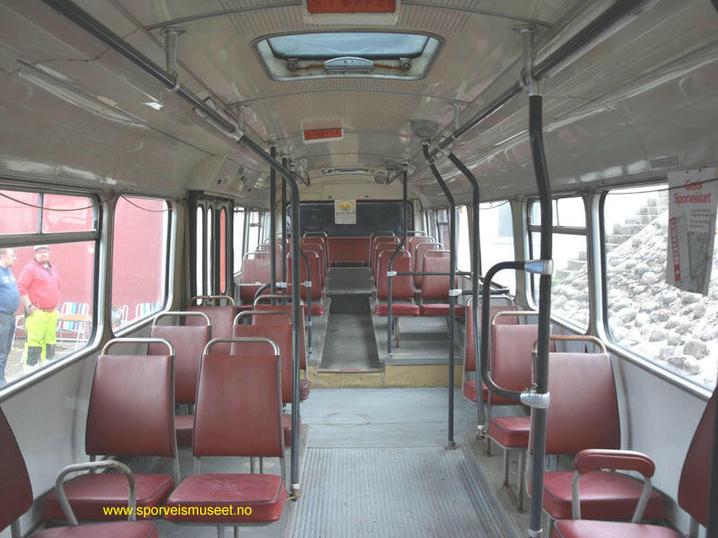 Buss med rødt eksteriør og to doble dører. Rundt bussen går en hvit og sort stripe. Bussens interiør består av lyst grått gulv og lyse tak med vinrøde seter. 
