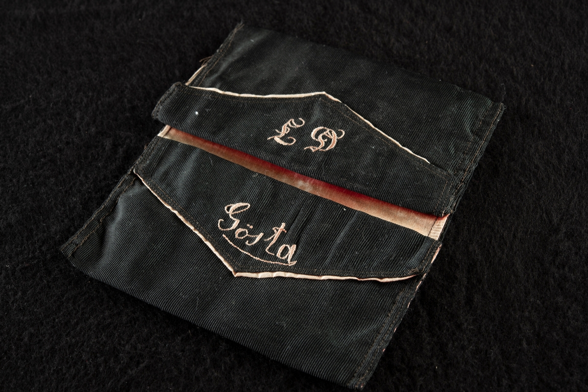 Plånbok av svart ripssiden. Invändigt klädd med rött siden. Broderad i rött med "Gösta" och "G. F." Det röda fodret samt bokstäverna har blekts betydligt och har nu en blekt gul färg.