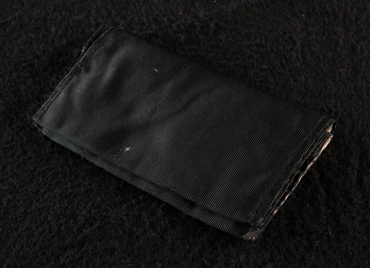 Plånbok av svart ripssiden. Invändigt klädd med rött siden. Broderad i rött med "Gösta" och "G. F." Det röda fodret samt bokstäverna har blekts betydligt och har nu en blekt gul färg.