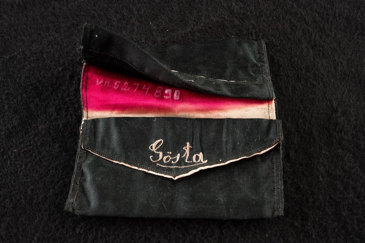Plånbok av svart ripssiden. Invändigt klädd med rött siden. Broderad i rött med "Gösta" och "G. F." Det röda fodret samt bokstäverna har blekts betydligt och har nu en blekt gul färg.