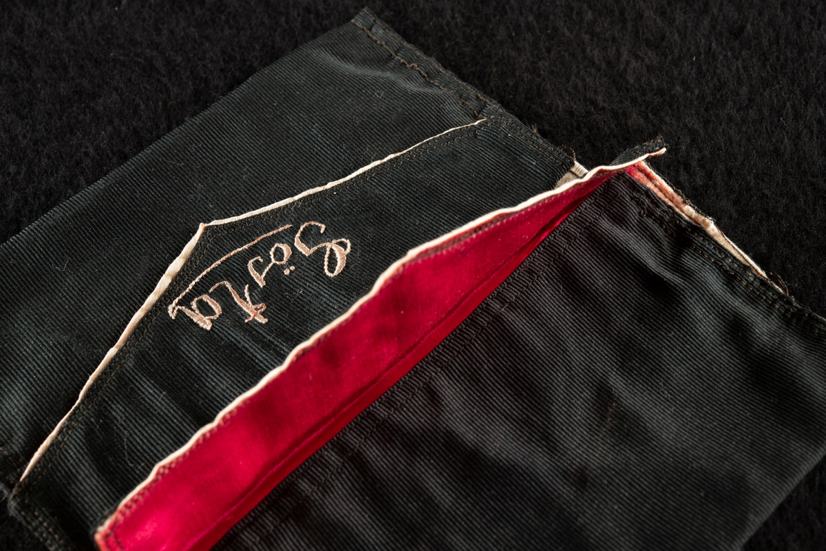 Plånbok av svart ripssiden. Invändigt klädd med rött siden. Broderad i rött med "Gösta" och "G. F." Det röda fodret samt bokstäverna har blekts betydligt och har nu en blekt gul färg.