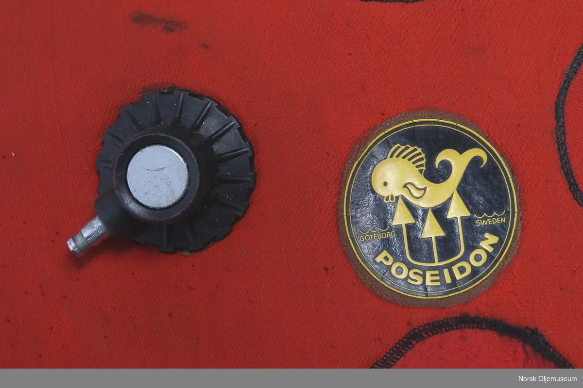 Tørrdrakt av neopren. Dykkerdrakten er av merke Poseidon og det er utført reparasjoner og endringer på drakten. Den har påsatte svarte sko som ikke er originale og utventil er plassert øverst på venstre arm. Foran på brystet har den to påsydde runde lapper av neopren. Drakten har slitasje på knærne fra bruk.