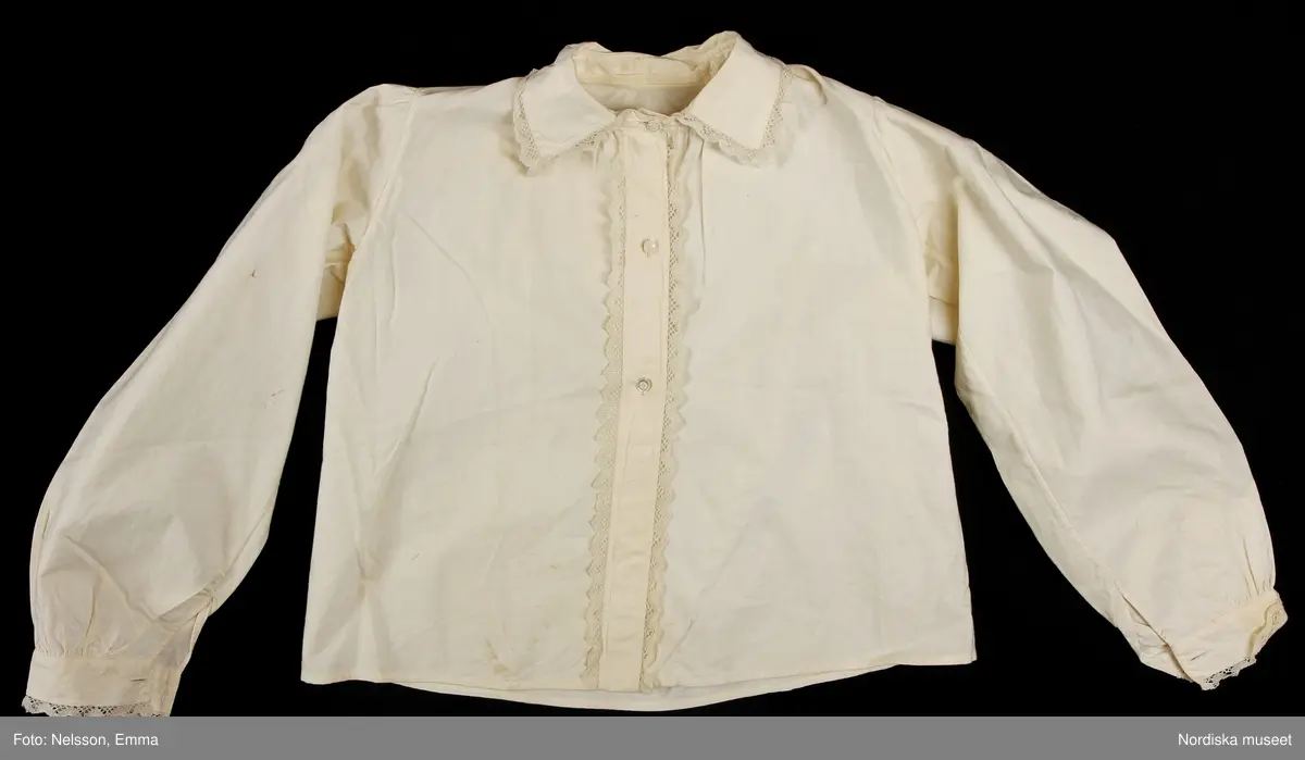 Katalogkort:
""Österåkersdräkt", 5 delar, för 8-åring. 
Bestående av överdel, livstyckekjol, förkläde, huvudbonad och bälte. 
Överdelen av vit bomullslärft med knypplad kantspets. 
Livstyckekjolen; rött kläde i kjolen, livet av gul sidenatlas med silkebroderier.
Förklädet av grön kläde med bårder nedtill i silkebroderier samt bandgarnering. Midjelinningen samt kantslå nedtill av svart kläde även dessa broderade. 
Bälte av rött läder med silkebroderier. 
Huvudbonad, best. av röda valkar, spetsluva och tyllhuckle."