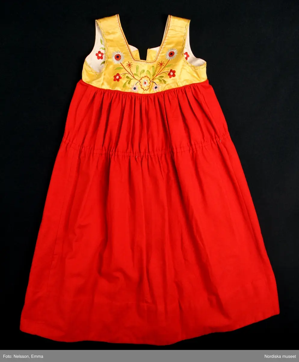 Katalogkort:
""Österåkersdräkt", 5 delar, för 8-åring. 
Bestående av överdel, livstyckekjol, förkläde, huvudbonad och bälte. 
Överdelen av vit bomullslärft med knypplad kantspets. 
Livstyckekjolen; rött kläde i kjolen, livet av gul sidenatlas med silkebroderier.
Förklädet av grön kläde med bårder nedtill i silkebroderier samt bandgarnering. Midjelinningen samt kantslå nedtill av svart kläde även dessa broderade. 
Bälte av rött läder med silkebroderier. 
Huvudbonad, best. av röda valkar, spetsluva och tyllhuckle."