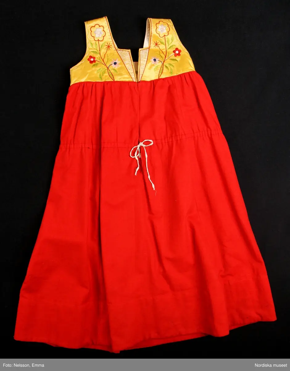 Katalogkort:
""Österåkersdräkt", 5 delar, för 8-åring. 
Bestående av överdel, livstyckekjol, förkläde, huvudbonad och bälte. 
Överdelen av vit bomullslärft med knypplad kantspets. 
Livstyckekjolen; rött kläde i kjolen, livet av gul sidenatlas med silkebroderier.
Förklädet av grön kläde med bårder nedtill i silkebroderier samt bandgarnering. Midjelinningen samt kantslå nedtill av svart kläde även dessa broderade. 
Bälte av rött läder med silkebroderier. 
Huvudbonad, best. av röda valkar, spetsluva och tyllhuckle."