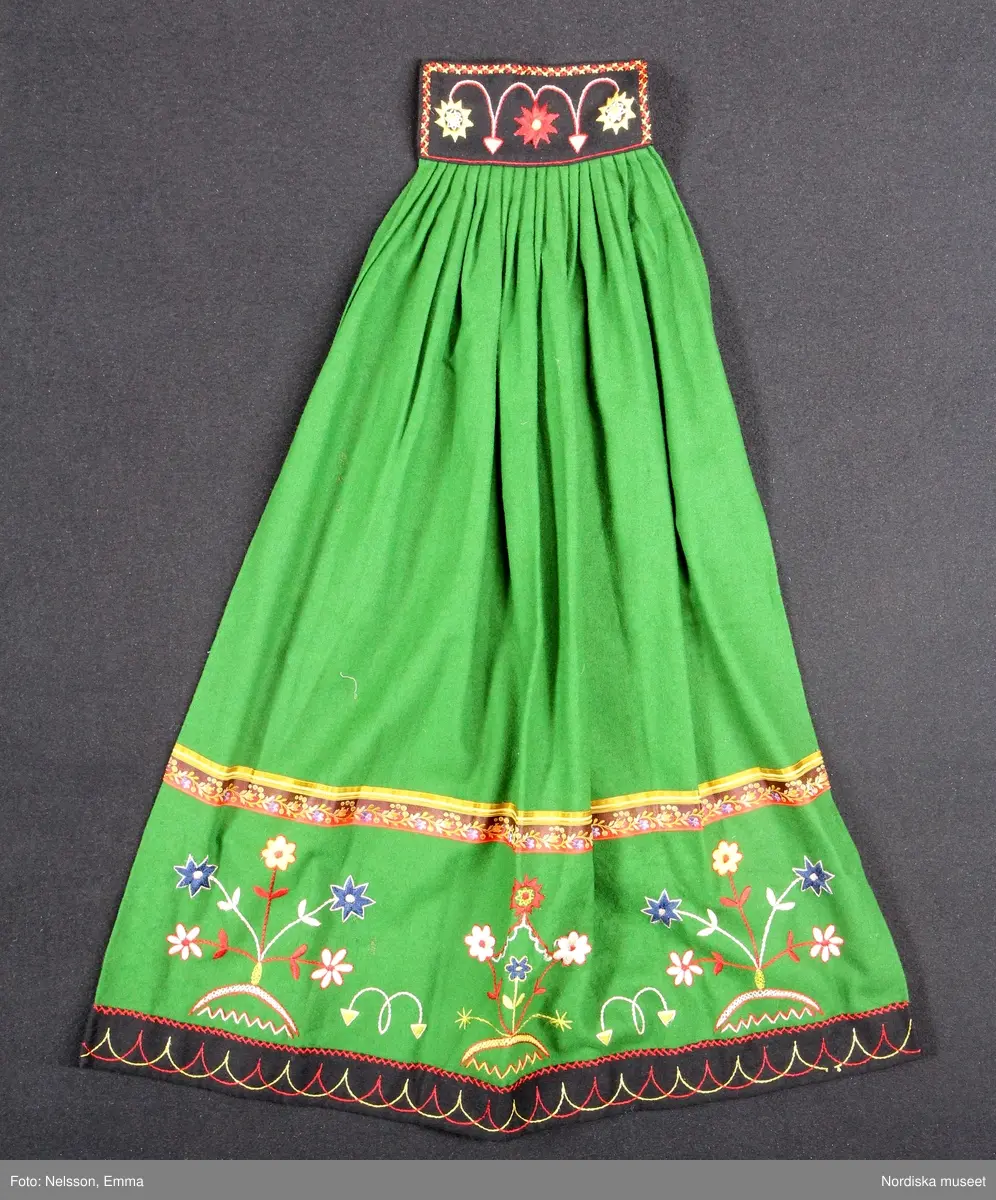 Katalogkort:
""Österåkersdräkt", 5 delar, för 8-åring. 
Bestående av överdel, livstyckekjol, förkläde, huvudbonad och bälte. 
Överdelen av vit bomullslärft med knypplad kantspets. 
Livstyckekjolen; rött kläde i kjolen, livet av gul sidenatlas med silkebroderier.
Förklädet av grön kläde med bårder nedtill i silkebroderier samt bandgarnering. Midjelinningen samt kantslå nedtill av svart kläde även dessa broderade. 
Bälte av rött läder med silkebroderier. 
Huvudbonad, best. av röda valkar, spetsluva och tyllhuckle."