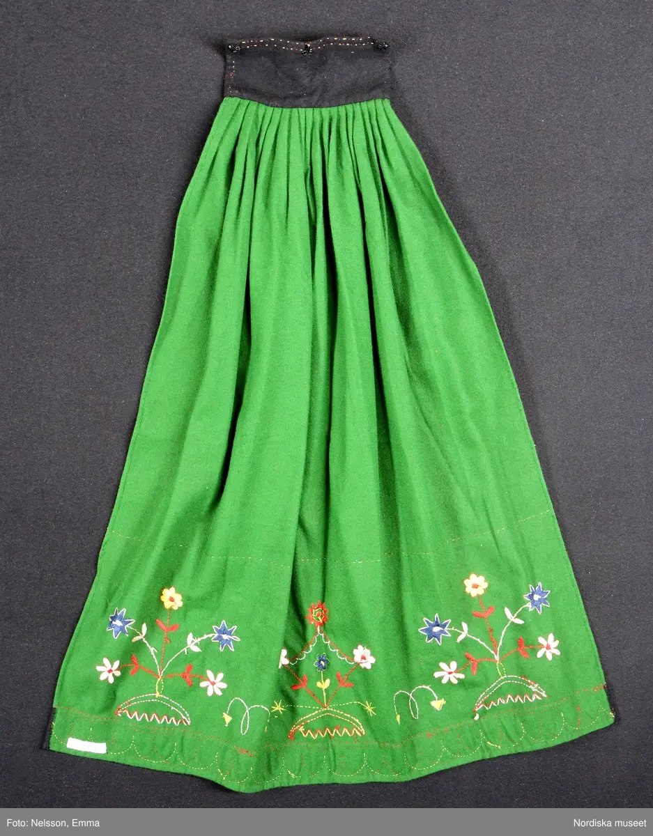 Katalogkort:
""Österåkersdräkt", 5 delar, för 8-åring. 
Bestående av överdel, livstyckekjol, förkläde, huvudbonad och bälte. 
Överdelen av vit bomullslärft med knypplad kantspets. 
Livstyckekjolen; rött kläde i kjolen, livet av gul sidenatlas med silkebroderier.
Förklädet av grön kläde med bårder nedtill i silkebroderier samt bandgarnering. Midjelinningen samt kantslå nedtill av svart kläde även dessa broderade. 
Bälte av rött läder med silkebroderier. 
Huvudbonad, best. av röda valkar, spetsluva och tyllhuckle."