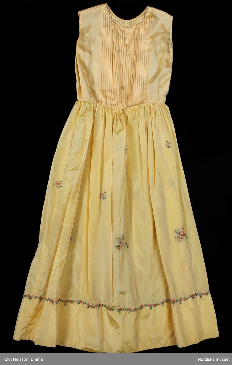 Katalogkort:
"Klänning med bolero för tonårsflicka av gul konstsidentaft (livet i något ljusare nyans) m. broderi i form av strödda blommor + bård nedtill i rosa, blått o. grönt brodérsilke.

Klänning. Avskuren i livet. Ärmlös. Livet sytt av 1 fram- o. 1 bakstycke. Plisserat mittparti både fram o. bak. Rundringad hals m. slå av tyget. Knäppning vid halsen o. i midjan bak m. tryckknappar. Kjolen består av 1 fram- o. 1 bakvåd, rynkad mot midjan. Ärmlappar av vit organza, vars gummering har torkat. En 15 cm bred skoning nedtill på kjolens insida. 

Bolero. Bestående av 2 framstycken + 1 bakstycke. Isydda korta puffärmar. Framkanten rundad nedtill. Framflyttad axelsöm. Midjeinsyningar.

Tillhörande skärp av sidenrips i samma nyans som klänningen. Överklätt rektangulärt spänne. Skärpänden m. tryckknapp."