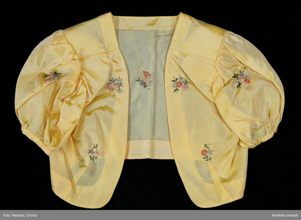 Katalogkort:
"Klänning med bolero för tonårsflicka av gul konstsidentaft (livet i något ljusare nyans) m. broderi i form av strödda blommor + bård nedtill i rosa, blått o. grönt brodérsilke.

Klänning. Avskuren i livet. Ärmlös. Livet sytt av 1 fram- o. 1 bakstycke. Plisserat mittparti både fram o. bak. Rundringad hals m. slå av tyget. Knäppning vid halsen o. i midjan bak m. tryckknappar. Kjolen består av 1 fram- o. 1 bakvåd, rynkad mot midjan. Ärmlappar av vit organza, vars gummering har torkat. En 15 cm bred skoning nedtill på kjolens insida. 

Bolero. Bestående av 2 framstycken + 1 bakstycke. Isydda korta puffärmar. Framkanten rundad nedtill. Framflyttad axelsöm. Midjeinsyningar.

Tillhörande skärp av sidenrips i samma nyans som klänningen. Överklätt rektangulärt spänne. Skärpänden m. tryckknapp."