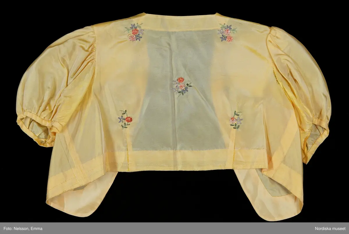 Katalogkort:
"Klänning med bolero för tonårsflicka av gul konstsidentaft (livet i något ljusare nyans) m. broderi i form av strödda blommor + bård nedtill i rosa, blått o. grönt brodérsilke.

Klänning. Avskuren i livet. Ärmlös. Livet sytt av 1 fram- o. 1 bakstycke. Plisserat mittparti både fram o. bak. Rundringad hals m. slå av tyget. Knäppning vid halsen o. i midjan bak m. tryckknappar. Kjolen består av 1 fram- o. 1 bakvåd, rynkad mot midjan. Ärmlappar av vit organza, vars gummering har torkat. En 15 cm bred skoning nedtill på kjolens insida. 

Bolero. Bestående av 2 framstycken + 1 bakstycke. Isydda korta puffärmar. Framkanten rundad nedtill. Framflyttad axelsöm. Midjeinsyningar.

Tillhörande skärp av sidenrips i samma nyans som klänningen. Överklätt rektangulärt spänne. Skärpänden m. tryckknapp."