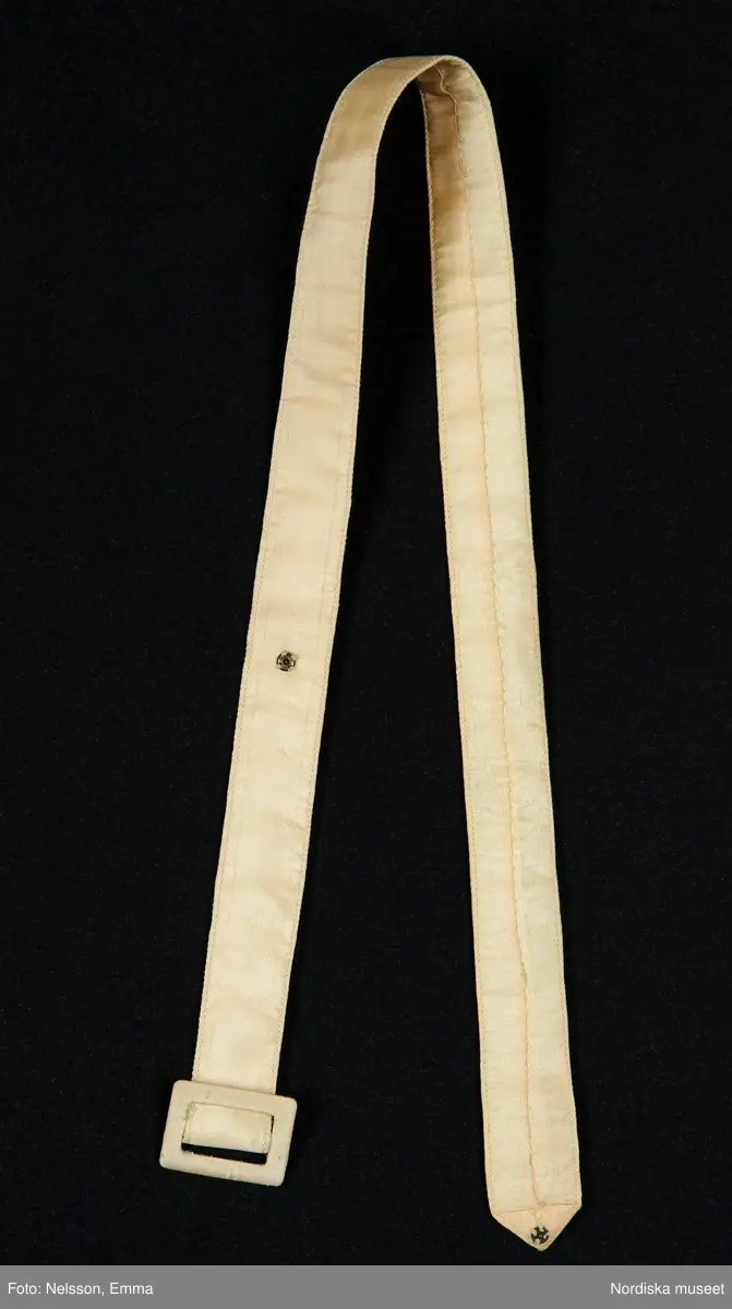 Katalogkort:
"Klänning med bolero för tonårsflicka av gul konstsidentaft (livet i något ljusare nyans) m. broderi i form av strödda blommor + bård nedtill i rosa, blått o. grönt brodérsilke.

Klänning. Avskuren i livet. Ärmlös. Livet sytt av 1 fram- o. 1 bakstycke. Plisserat mittparti både fram o. bak. Rundringad hals m. slå av tyget. Knäppning vid halsen o. i midjan bak m. tryckknappar. Kjolen består av 1 fram- o. 1 bakvåd, rynkad mot midjan. Ärmlappar av vit organza, vars gummering har torkat. En 15 cm bred skoning nedtill på kjolens insida. 

Bolero. Bestående av 2 framstycken + 1 bakstycke. Isydda korta puffärmar. Framkanten rundad nedtill. Framflyttad axelsöm. Midjeinsyningar.

Tillhörande skärp av sidenrips i samma nyans som klänningen. Överklätt rektangulärt spänne. Skärpänden m. tryckknapp."