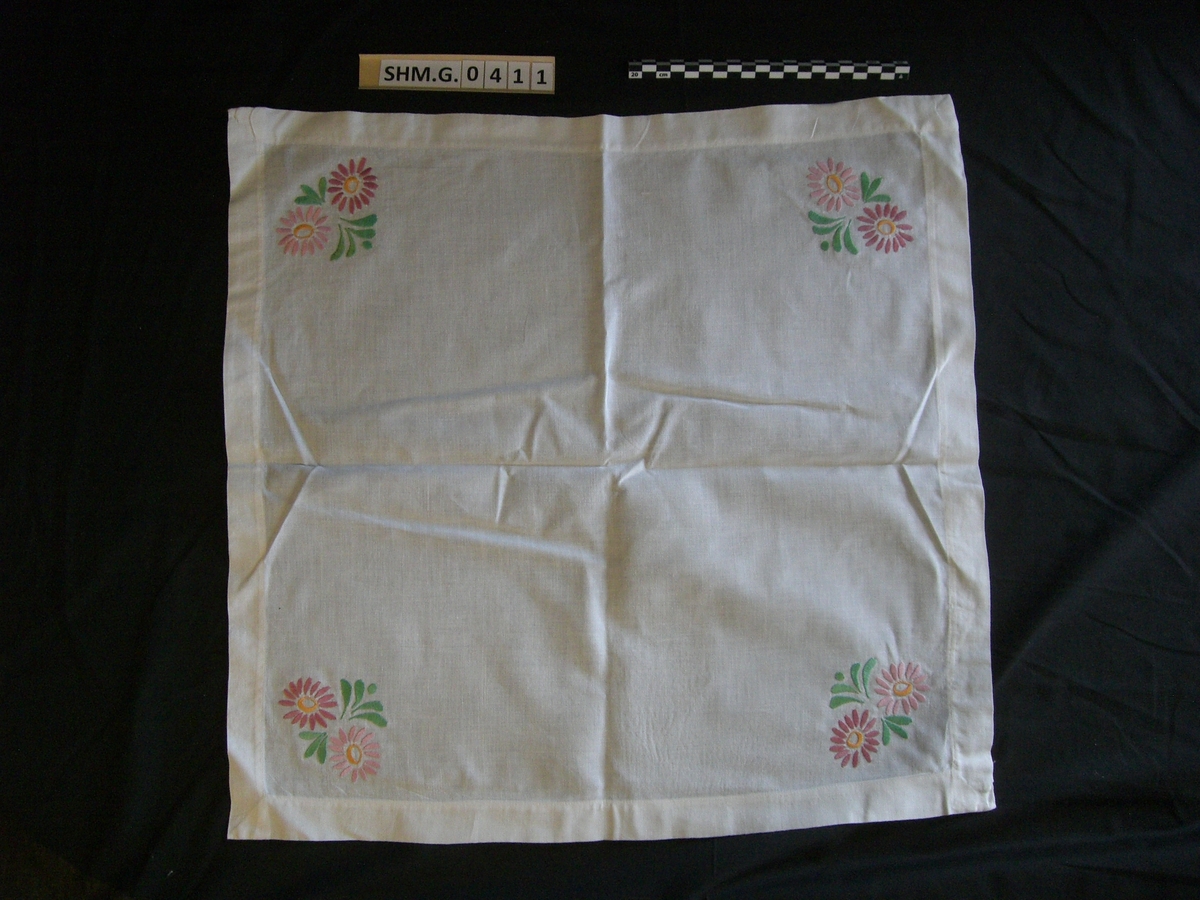 Hvit duk i cambic med 3 cm bred maskinsydd fald. Brodert (plattsaum) blomstermotiv i moulinegarn i hjørnene. Motiv i grønt, gult og 2 lilla nyanser.