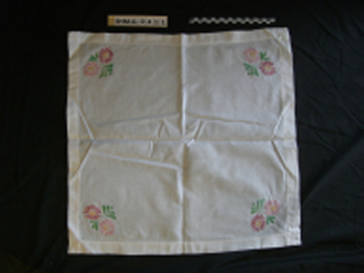 Hvit duk i cambic med 3 cm bred maskinsydd fald. Brodert (plattsaum) blomstermotiv i moulinegarn i hjørnene. Motiv i grønt, gult og 2 lilla nyanser.
