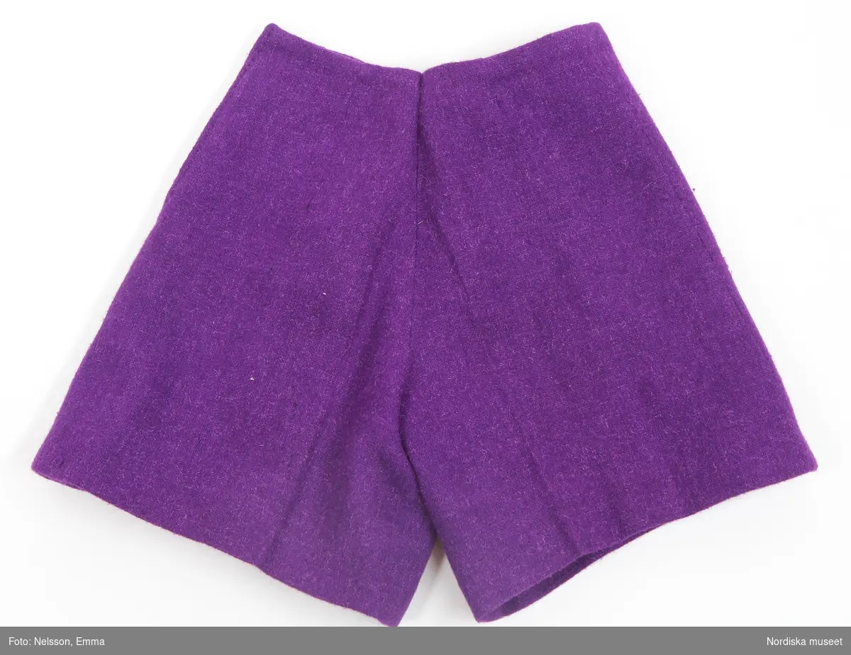 Katalogkort:
"Byxdräkt för flicka av starkt violett tunn ylletweed, lårkorta byxor s.k. "hot pants" och kortkort kappa som gå strax nedanför byxorna. 
Kappa: 2 framstycken med insprovning i midjan, 1 bakstycke, isydd lång ärm, ståndkrage som knäpps med en flik vid sidan, enkelknäppt högt i halsen med 7 överklädda knappar, 2 påsydda bröstfickor med motveck och ficklock med likadana klädda knappar, fodrad med violett konstsiden.
Skärp: löst skärp med stort tygklätt spänne och 6 nitade hå, samma foder som kappan.
Byxor: 2 fram- och 2 bakstycken, raka korta ben, linning saknas, blixtlås i vänster sidsöm, ofodrade.