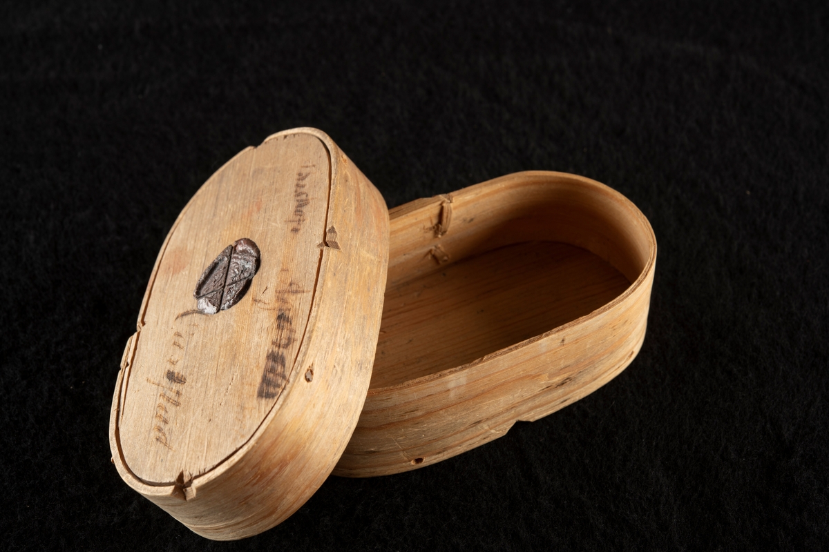 Oval, av träspån. På lock och botten kvarsittande lack, som varit genomdragna av snören.