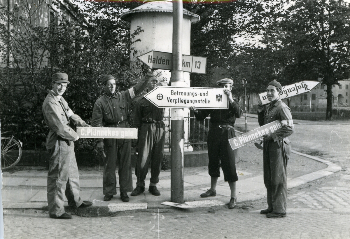 Fredrikstad. Vestsiden. Phønixplassen. Ferjestedsveien/J. N. Jacobsens gate. Biblioteket i bakgrunnen. Hjemmefronten. Henvisnings- og gatenavn- skilt fjernes. (Gatenavnskiltet henspiller på C. Plünnekes gate, som gikk mellom Nygaardsgaten og Storgaten, nåværende Doktor Giebelhausens gate). Carl Plünneke var opprinnelig en tysk barberer med stor interesse for Fredrikstads histore. Han utga en utall artikler og flere bøker om temaet. Under Den andre verdenskrig ble han beskyldt for å være tyskvennlig, og formannskapet beordret derfor på et møte mai/juni 1945 at gatenavnet skulle fjernes).