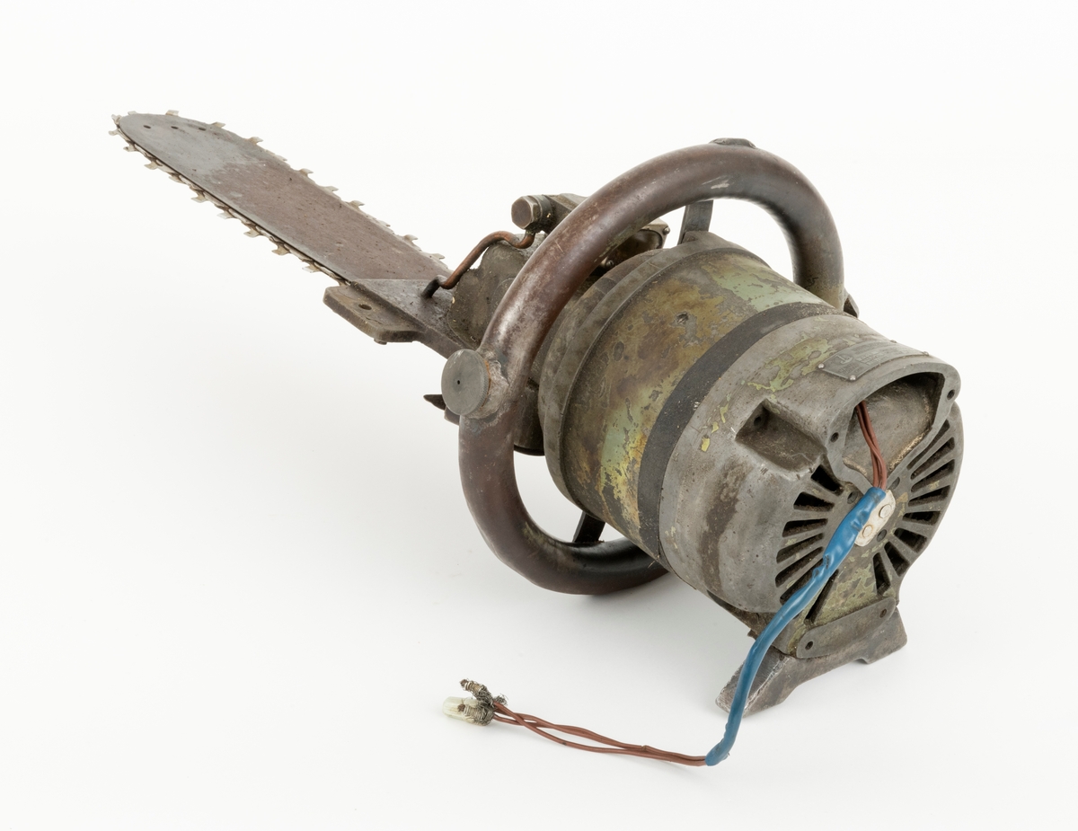 Motorsag med elektrisk drift.  Sager av denne typen ble mest brukt til kapping av virke på sagtomter.  Saga har rund motor med kombinert handtak og oljetank som ligger som et sirkulært rør rundt motoren. Motoren har rester av grønn lakk. Rundt motoren er det påsatt et tjæreband. Til saga er det koplet en 38 centimeter lang ledning. 