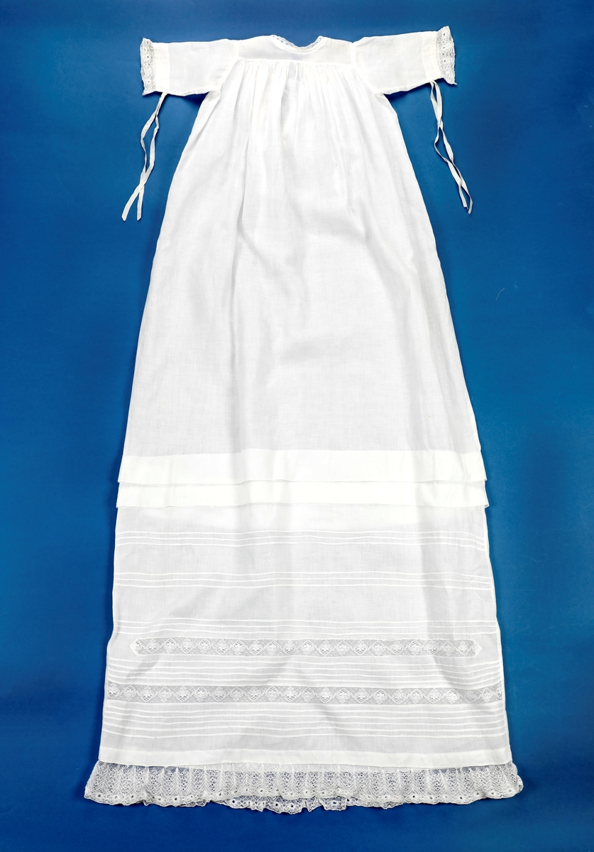 Lang dåpskjole med belte og underskjørt.
Kjolen har bærestykke hvor selve kjolen r rynket til. Halsringing, mansjetter og nedkant av skjørtet er besatt med blonder. Løpegang i halsen med knyting bak. Løpegang med knytesnor nederst på ermene. Splitt bak med to punkter med knytebånd. Forsiden av skjørtet er pyntet med biser og blonder.
Beltet er bredt, pyntet med biser og blonder i endene.
Underskjørtet har løpegang i livet og påsydd kappe i engelsk blonde.