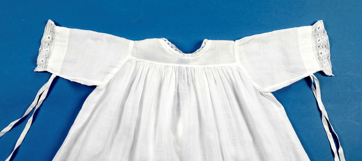 Lang dåpskjole med belte og underskjørt.
Kjolen har bærestykke hvor selve kjolen r rynket til. Halsringing, mansjetter og nedkant av skjørtet er besatt med blonder. Løpegang i halsen med knyting bak. Løpegang med knytesnor nederst på ermene. Splitt bak med to punkter med knytebånd. Forsiden av skjørtet er pyntet med biser og blonder.
Beltet er bredt, pyntet med biser og blonder i endene.
Underskjørtet har løpegang i livet og påsydd kappe i engelsk blonde.