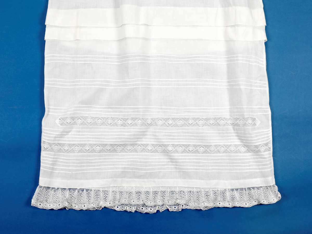 Lang dåpskjole med belte og underskjørt.
Kjolen har bærestykke hvor selve kjolen r rynket til. Halsringing, mansjetter og nedkant av skjørtet er besatt med blonder. Løpegang i halsen med knyting bak. Løpegang med knytesnor nederst på ermene. Splitt bak med to punkter med knytebånd. Forsiden av skjørtet er pyntet med biser og blonder.
Beltet er bredt, pyntet med biser og blonder i endene.
Underskjørtet har løpegang i livet og påsydd kappe i engelsk blonde.