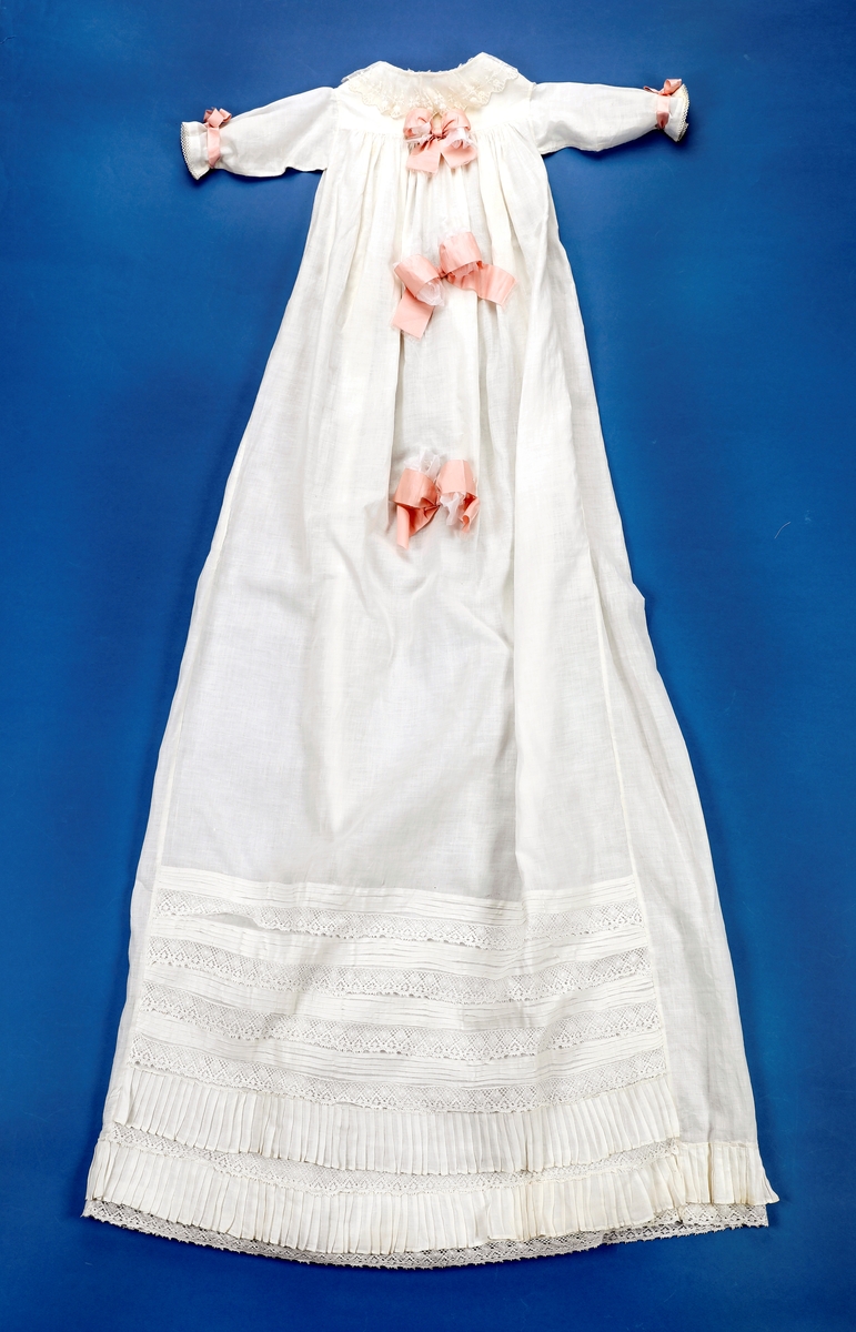 Dåpskjole med tilbehør: belte, lue, votter.  

Dåpskjole med isydde ermer, skulderstykke, langt vidt skjørt med rynker. Dekorteknikk: Foldelagte kapper 2 foran, 1 bak festet med blonde. Sydde legg, 5 stk., festet med blonde. Åpning bak 40 cm med 2 knytteband. pyntet med rosa bånd.
Lue:bobinet foret med rosa sateng. 
belte: bredt og langt med pynt av blonder og biser i hver ende.
Strikkede votter, tradisjonell form med knyttesnor.