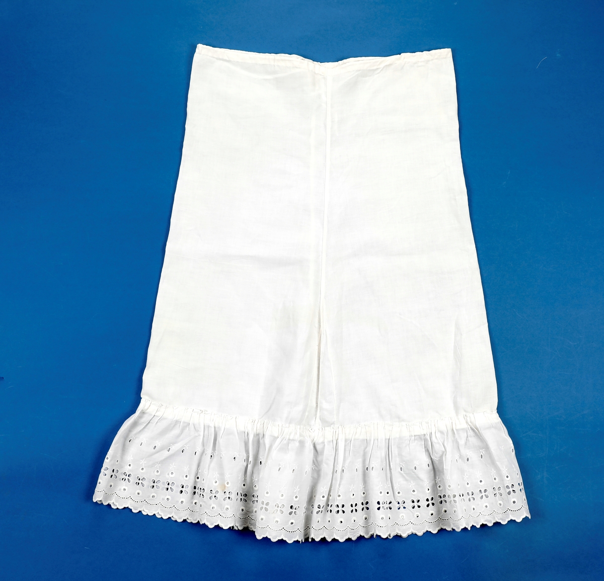 Lang dåpskjole med belte og underskjørt.
Kjolen har bærestykke hvor selve kjolen r rynket til. Halsringing, mansjetter og nedkant av skjørtet er besatt med blonder. Løpegang i halsen med knyting bak. Løpegang med knytesnor nederst på ermene. Splitt bak med to punkter med knytebånd. Forsiden av skjørtet er pyntet med biser og blonder.
Beltet er bredt, pyntet med biser og blonder i endene.
Underskjørtet har løpegang i livet og påsydd kappe i engelsk blonde.