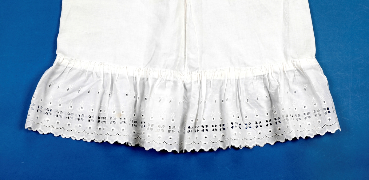 Lang dåpskjole med belte og underskjørt.
Kjolen har bærestykke hvor selve kjolen r rynket til. Halsringing, mansjetter og nedkant av skjørtet er besatt med blonder. Løpegang i halsen med knyting bak. Løpegang med knytesnor nederst på ermene. Splitt bak med to punkter med knytebånd. Forsiden av skjørtet er pyntet med biser og blonder.
Beltet er bredt, pyntet med biser og blonder i endene.
Underskjørtet har løpegang i livet og påsydd kappe i engelsk blonde.