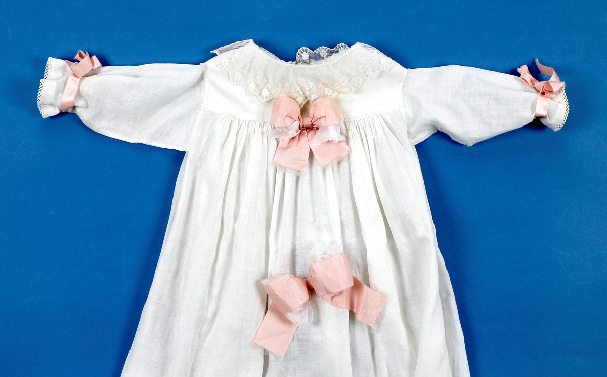 Dåpskjole med tilbehør: belte, lue, votter.  

Dåpskjole med isydde ermer, skulderstykke, langt vidt skjørt med rynker. Dekorteknikk: Foldelagte kapper 2 foran, 1 bak festet med blonde. Sydde legg, 5 stk., festet med blonde. Åpning bak 40 cm med 2 knytteband. pyntet med rosa bånd.
Lue:bobinet foret med rosa sateng. 
belte: bredt og langt med pynt av blonder og biser i hver ende.
Strikkede votter, tradisjonell form med knyttesnor.