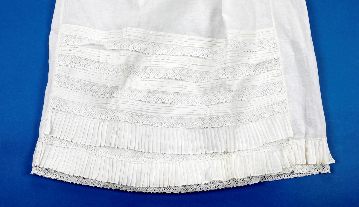 Dåpskjole med tilbehør: belte, lue, votter.  

Dåpskjole med isydde ermer, skulderstykke, langt vidt skjørt med rynker. Dekorteknikk: Foldelagte kapper 2 foran, 1 bak festet med blonde. Sydde legg, 5 stk., festet med blonde. Åpning bak 40 cm med 2 knytteband. pyntet med rosa bånd.
Lue:bobinet foret med rosa sateng. 
belte: bredt og langt med pynt av blonder og biser i hver ende.
Strikkede votter, tradisjonell form med knyttesnor.