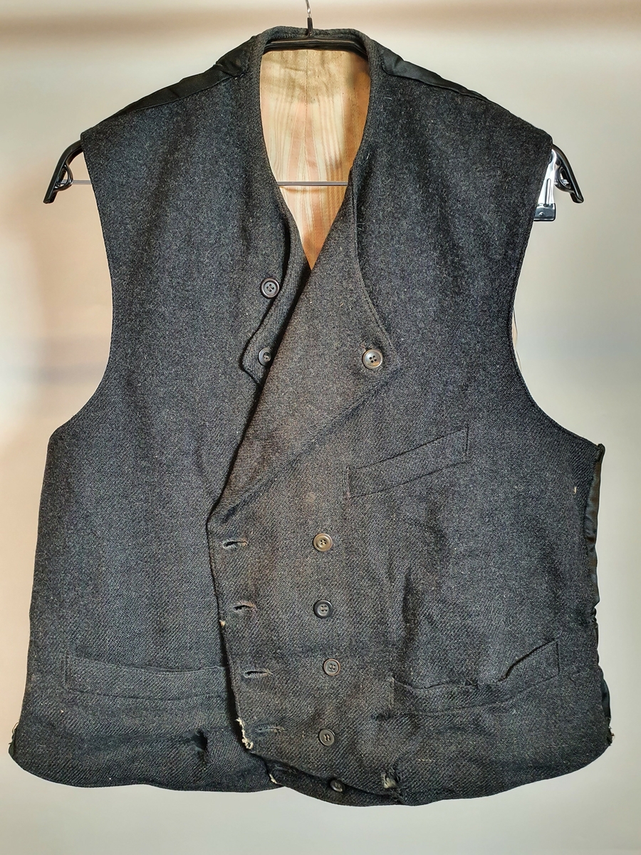 Sort vadmel.
Maskinsydd.
Dobbeltknappet med en knapp.
En ekstra knapp på hver side.
Tre lommer.
Ryggen sydd i silke.
Spensel med metallspenne.
Foret på forstykkene er i stripet dresstoff i sort og grått.
Lomme av bolster.
Foret i ryggen også bolster i rosa og hvit.
Slitt og hullet.