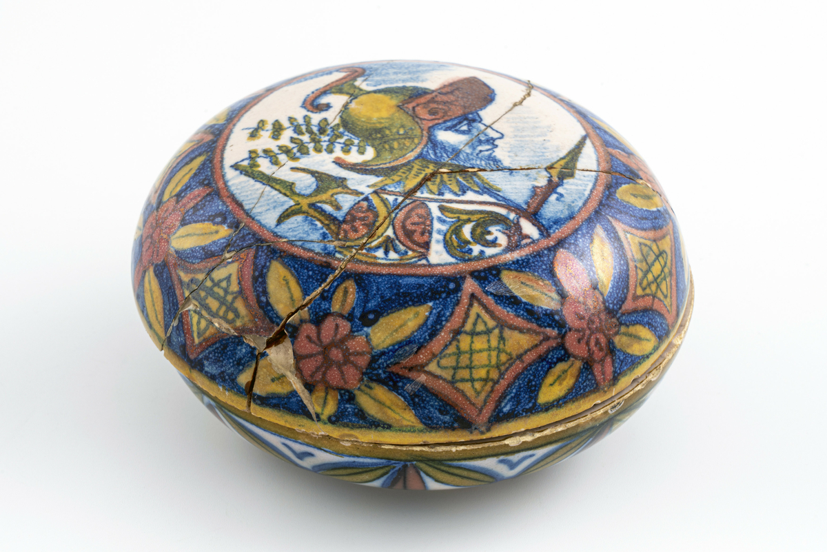 Liten keramikkskål med lokk, smaler mye av mot bunn og topp. Påmalt dekor med romersk (?) soldat i profil med hjelm, spyd og øks. Lokket har vært knust i flere biter, og er limt sammen igjen. Har tilhørt Sara Fabricius.