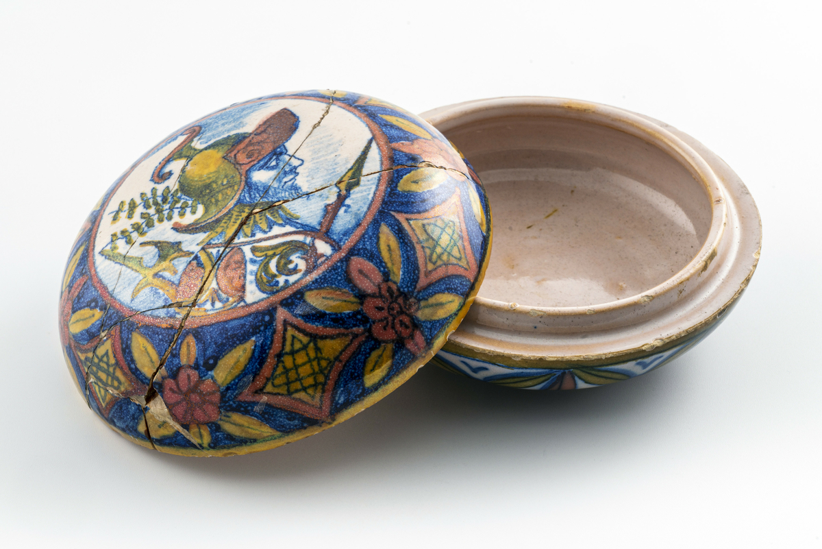 Liten keramikkskål med lokk, smaler mye av mot bunn og topp. Påmalt dekor med romersk (?) soldat i profil med hjelm, spyd og øks. Lokket har vært knust i flere biter, og er limt sammen igjen. Har tilhørt Sara Fabricius.