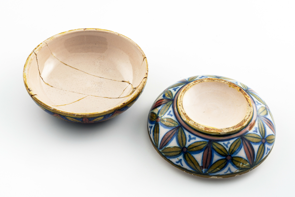 Liten keramikkskål med lokk, smaler mye av mot bunn og topp. Påmalt dekor med romersk (?) soldat i profil med hjelm, spyd og øks. Lokket har vært knust i flere biter, og er limt sammen igjen. Har tilhørt Sara Fabricius.